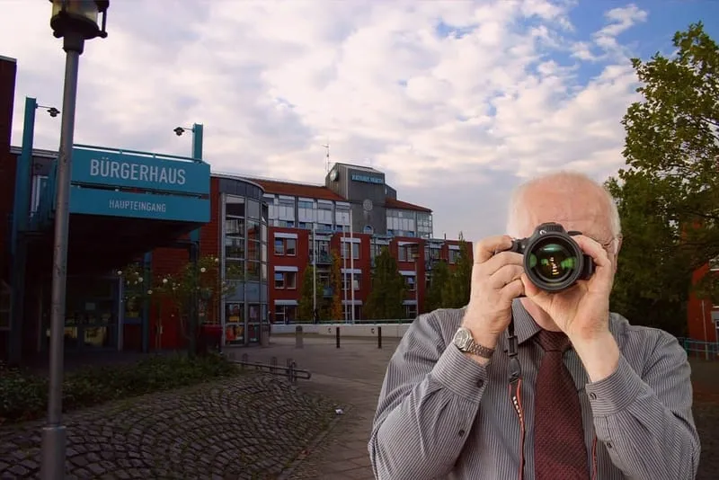 Detektiv in Hemd und Krawatte fotografiert vor Backsteingebäude mit Schild "Bürgerhaus Haupteingang" bei Wolkenhimmel.