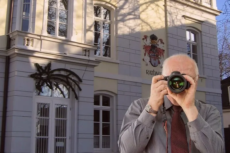 Detektiv mit Kamera in Hemd und Krawatte vor hellem Gebäude mit großen Fenstern, Wappen, Baumschatten - Detektei-Einsatz.