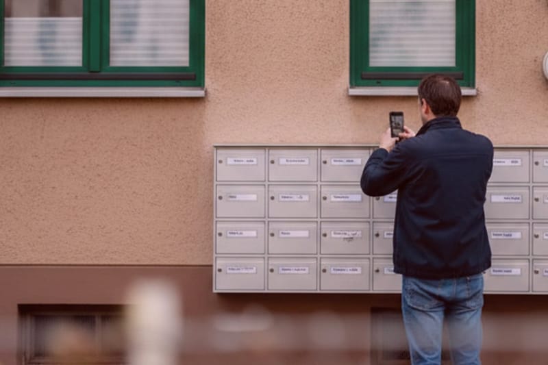 Detektiv fotografiert Briefkästen zur Adressermittlung außen an beiger Wand, Detektei-Einsatz, Ermittler bei Beweisaufnahme.