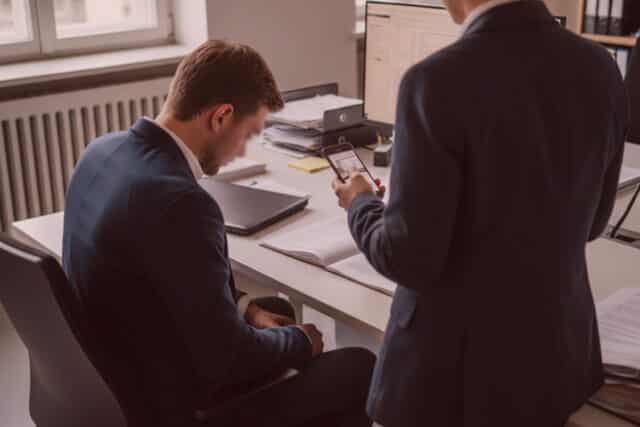 Detektive in Geschäftskleidung, einer sitzt an einem Schreibtisch und sieht entmutigt aus, ein anderer prüft Dokumente mit einem Smartphone im Büro der Agentur.