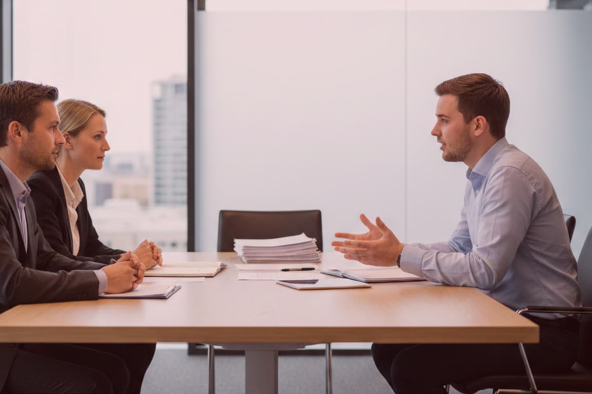 Detektive im modernen Büro bei einer Befragung: Zwei Ermittler mit Unterlagen, eine Person spricht gestikulierend gegenüber.