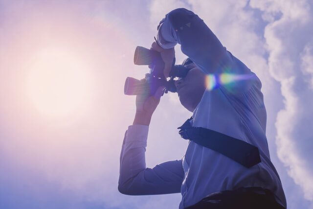 Detektei Ermittler beobachtet den Himmel mit Fernglas, Sonnenlicht und Lens Flare, professionelle Detektivarbeit im Einsatz.