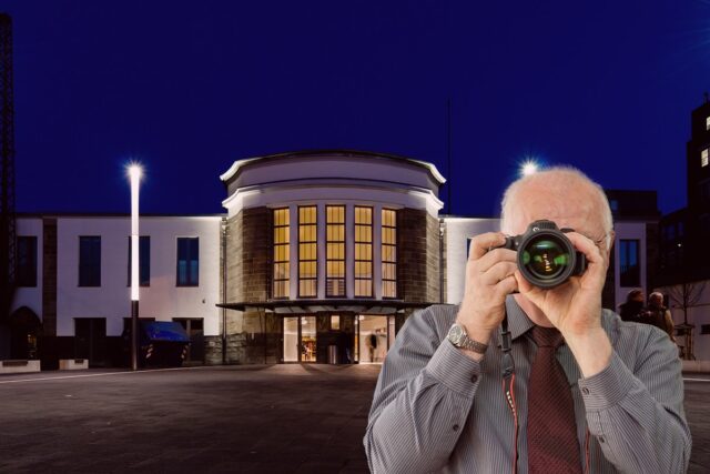 Detektei Ermittler fotografiert Beweise: Älterer Mann mit Kamera vor beleuchtetem Gebäude bei Observation für die Detektei.