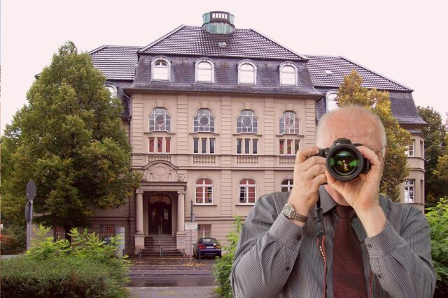 Erfahrener Detektiv fotografiert Beweismaterial vor historischem Gebäude mit Rundbogenfenstern und Bäumen - Detektei im Einsatz.