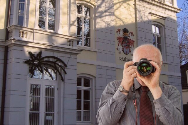 Detektiv mit Kamera in Hemd und Krawatte vor hellem Gebäude mit großen Fenstern, Wappen, Baumschatten - Detektei-Einsatz.
