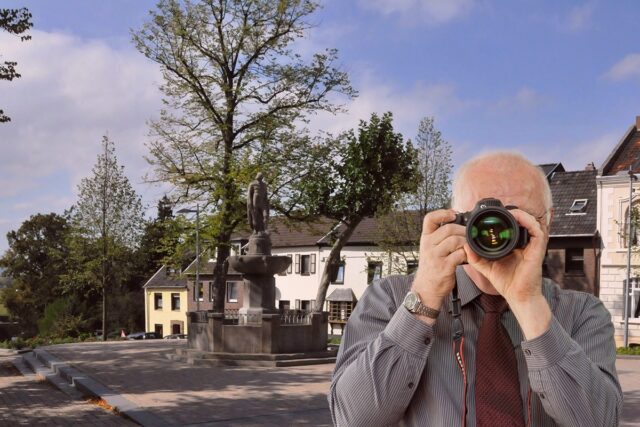 Ermittler fotografiert im Freien: Älterer Detektiv mit Kamera, Hemd und Krawatte, Statue und Häuser vor blauem Himmel.