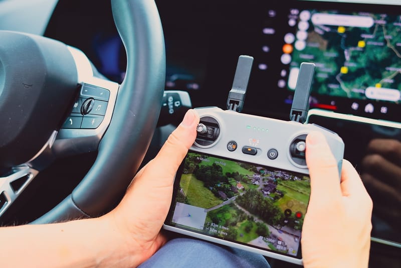 Detektiv steuert Drohne per Fernbedienung im Auto, überwacht Gelände auf Display, Lenkrad und Navigationsgerät sichtbar.