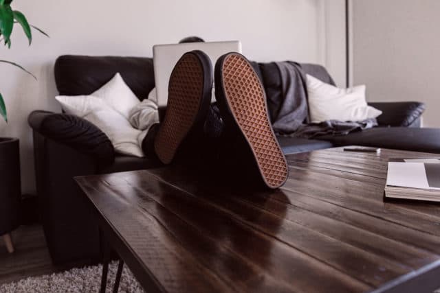 Kranker Mitarbeiter sitzt auf eine Couch mit einem Laptop, hat die Füße auf dem Tisch