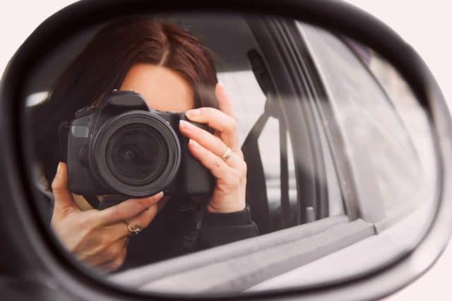 Privatdetektiv in einem Auto mit Kamera vor dem Gesicht durch einen Rückspiegel fotografiert
