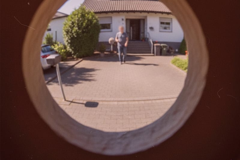 Detektiv überwacht Einfahrt durch Türspion: Person geht zur Haustür mit Ziegeldach, Auto links, unscharfes Bild.